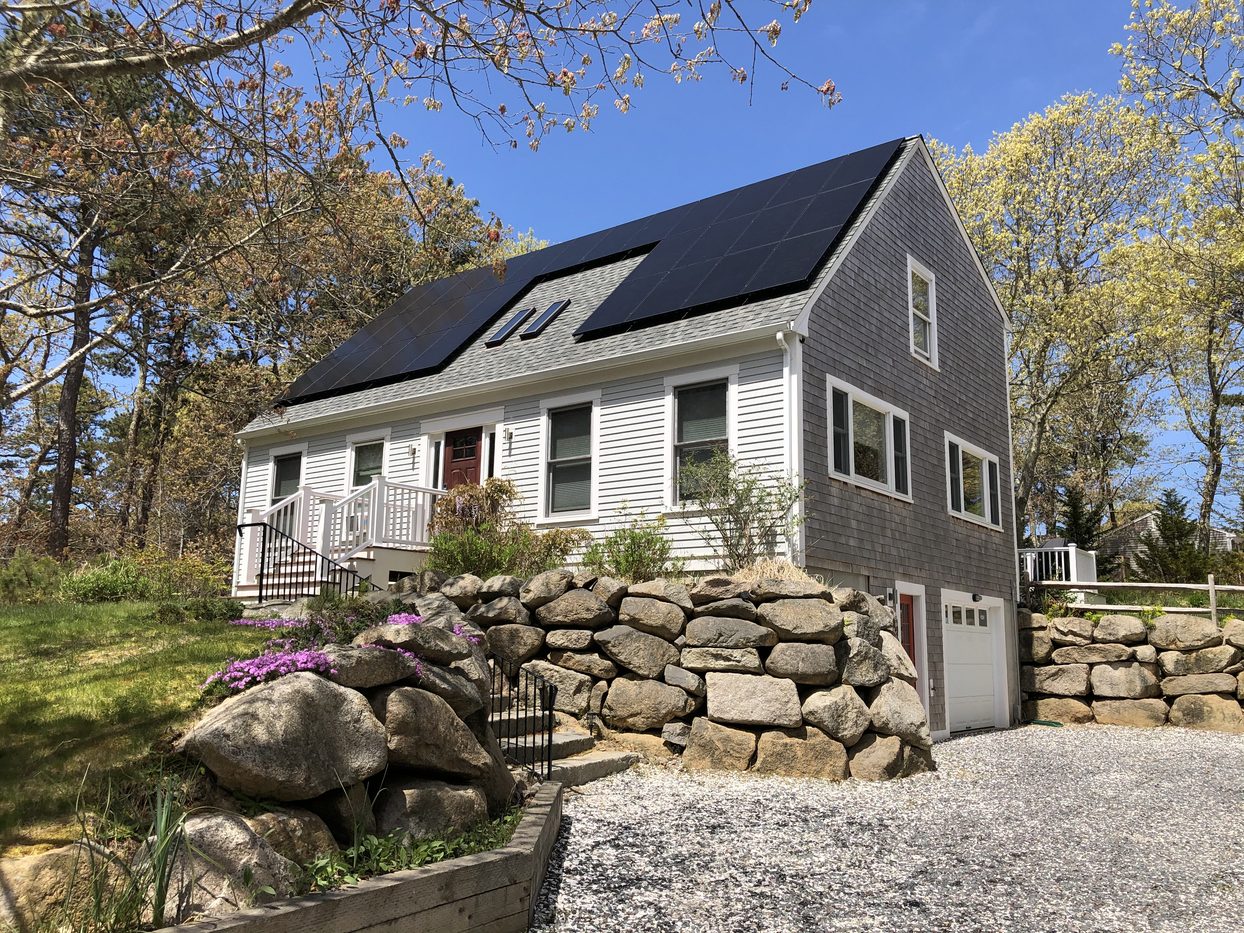 The Map House from the driveway: a gray-shingled Cape with solar panels above a stone wall